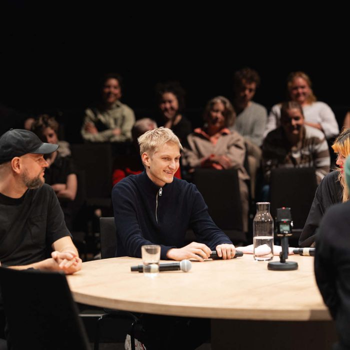 Ein junger Mann sitzt in der Mitte, er lächelt zufrieden links uns rechts sitzen zwei Personen, die ihm das Lächeln wohlwollend erwidern.